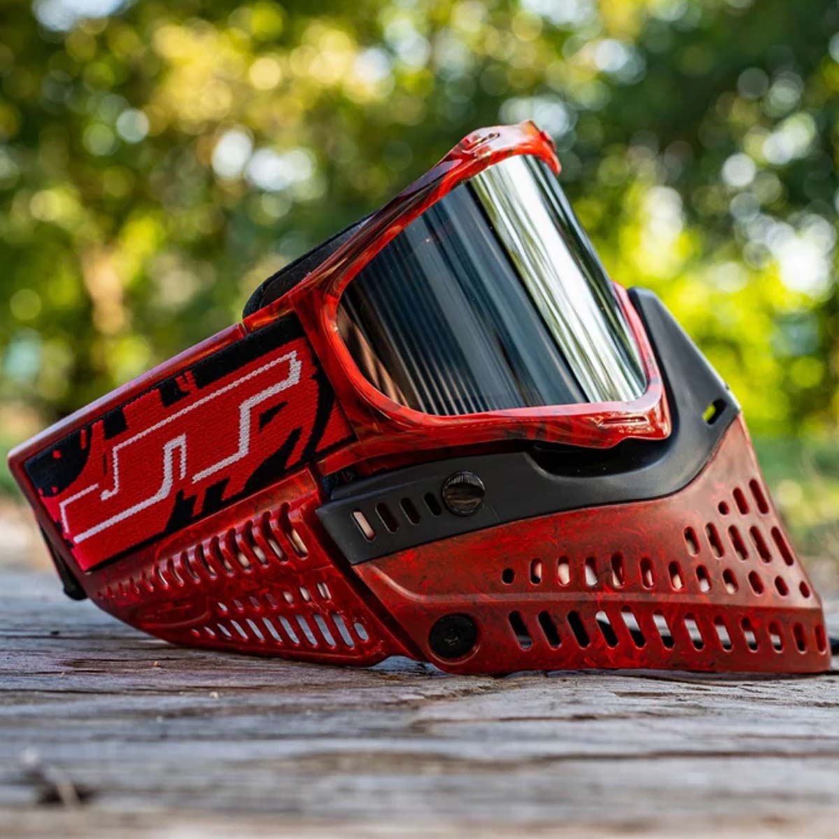 Red and black JT ProFlex Goggle LE Marble Red Black Home paintball mask with a smoke lens on a woodland background