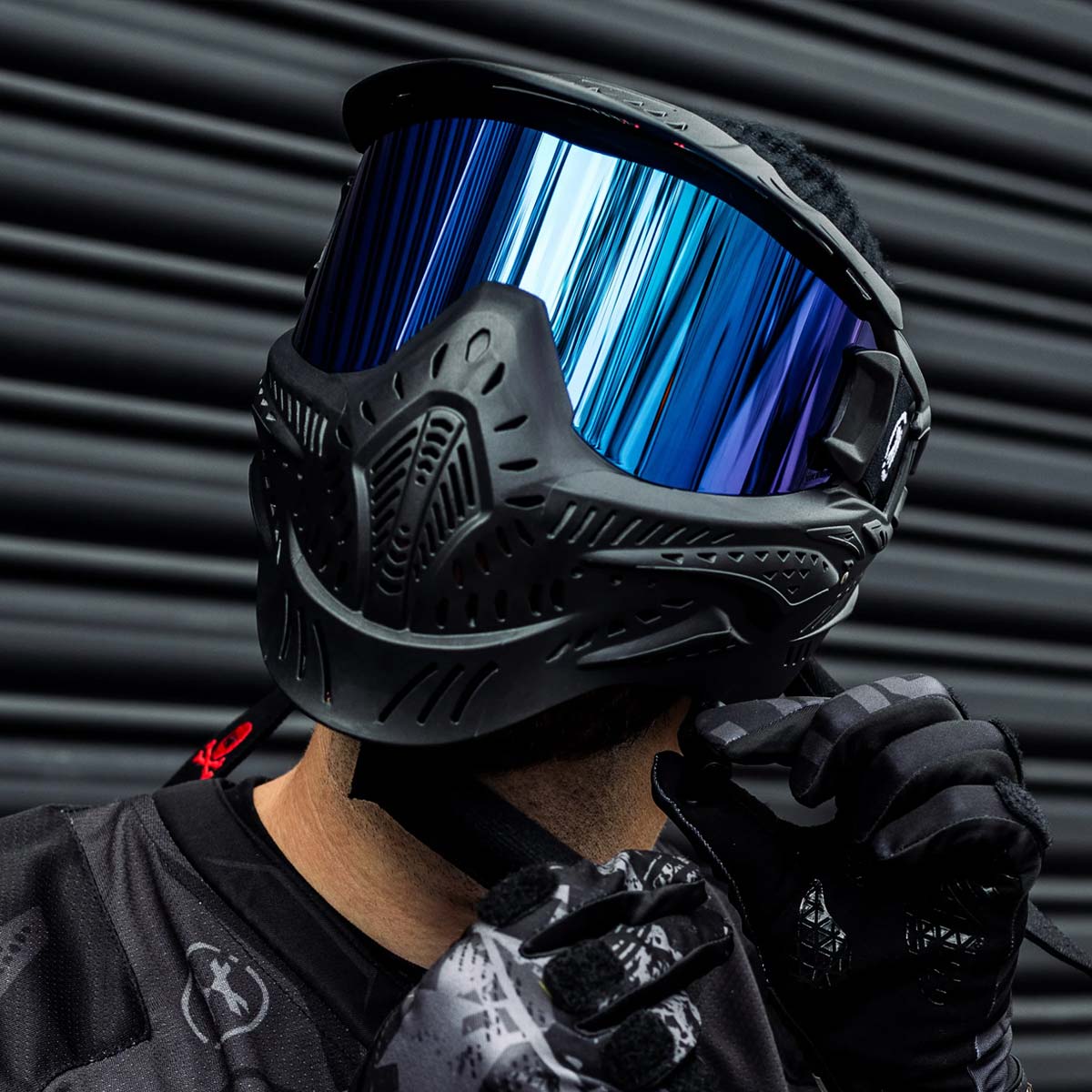 Black HK Army HSTL Thermal Paintball Goggle mask with a blue lens and white AK logo on the side, shown on a person fastening the chin strap