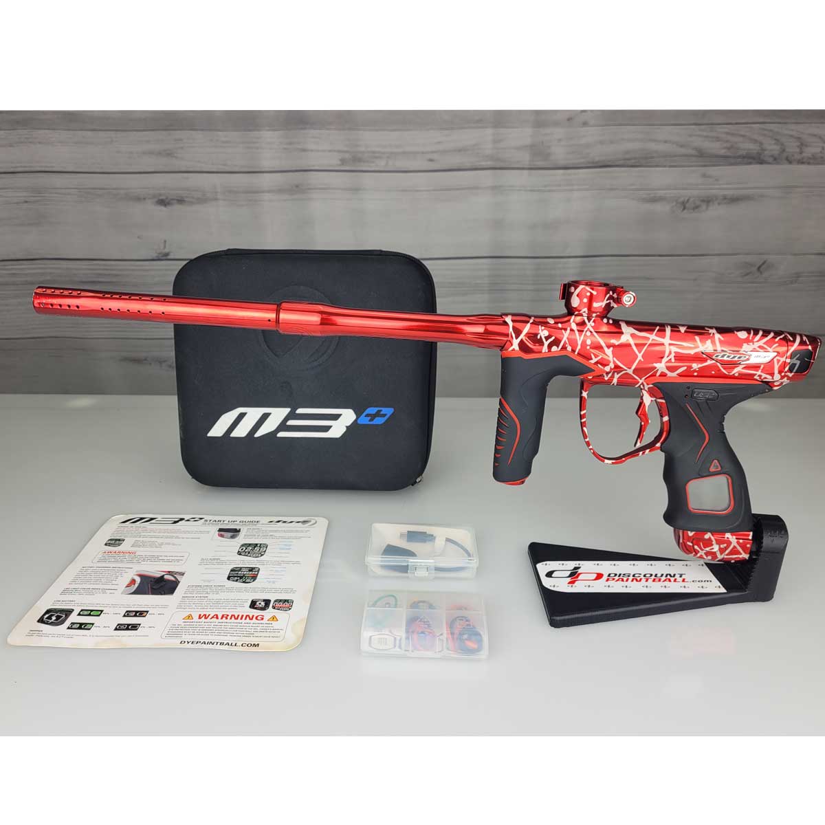 USED Red and black with white splash pattern Dye M3+ Paintball Marker Red Splash with case and spare parts kit on a woodgrain background.