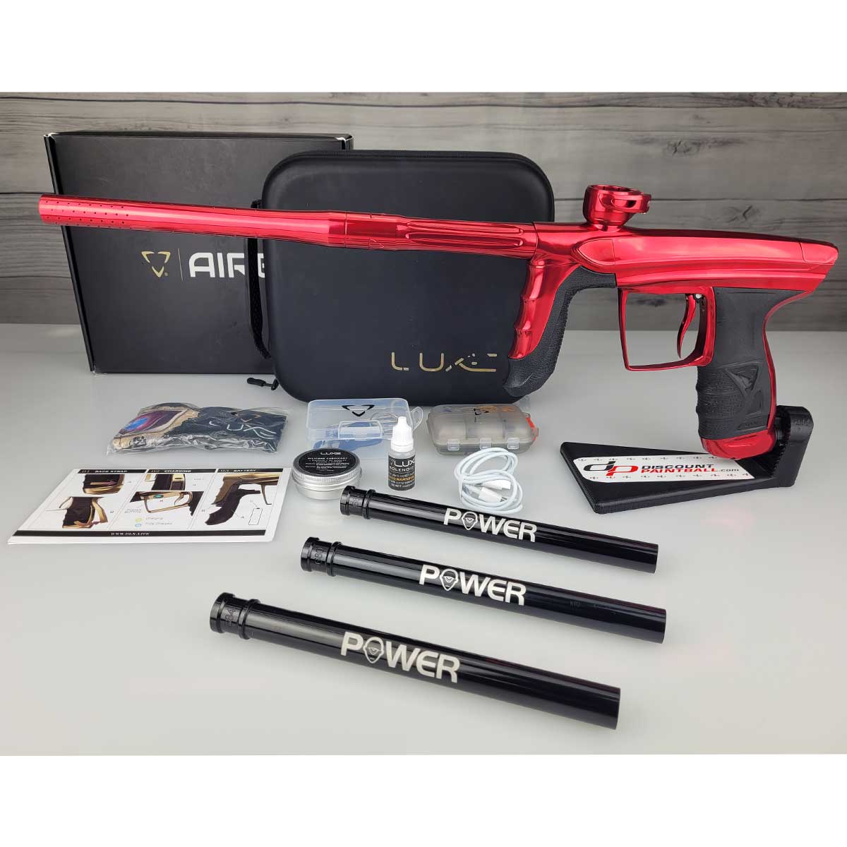 Left side USED DLX Luxe Aire Red Polished paintball marker on a stand with box, case, manual, barrel sock, 3 PWR inserts, tools and parts against a woodgrain background
