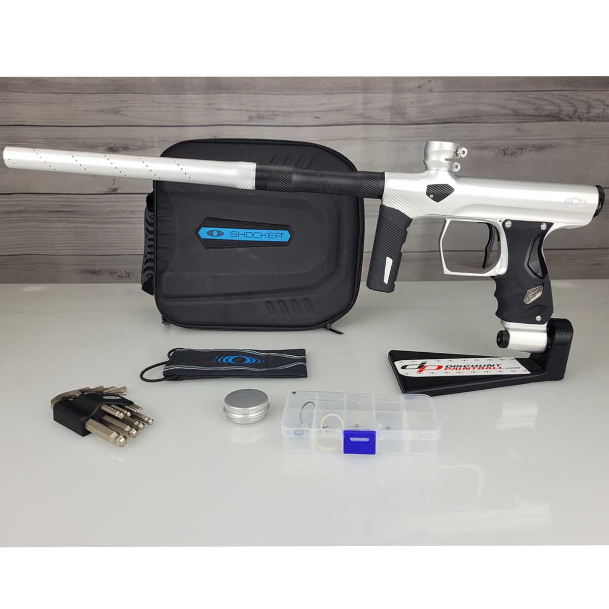 USED Shocker Era Silver black paintball marker in a stand with a case, tools, barrel sock, silver grease pot and clear parts kit on a woodgrain background.