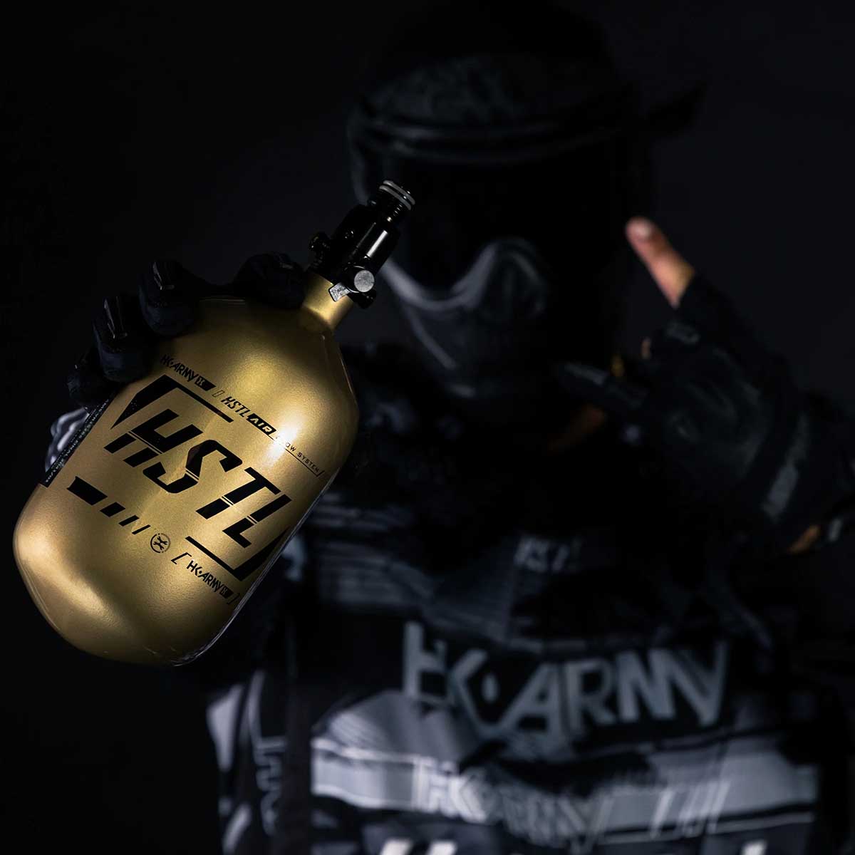 A player in HK Army clothing holding a HK Army HSTL 68ci 4500psi Carbon Fiber Air Tank Gold Black in one hand
