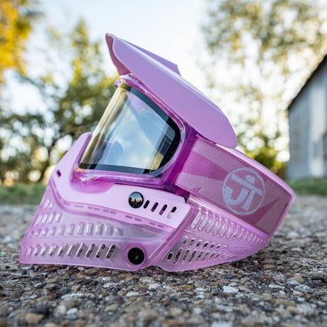Pink JT ProFlex Birthstone Ice October Opal Goggle paintball mask on a gravel surface with blurred greenery in the background