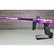 A USED DYE DSR Pro PGA Grimace Purple Splash paintball marker with a vibrant purple and black lightning-patterned finish, displayed on a black stand against a wood-plank background.
