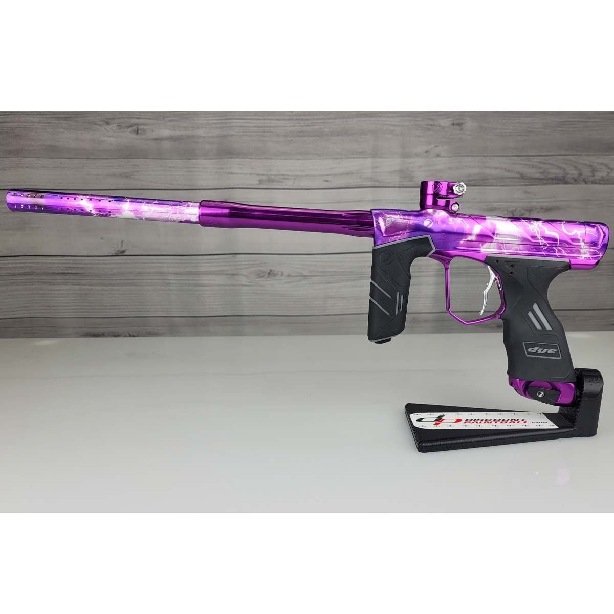 A USED DYE DSR Pro PGA Grimace Purple Splash paintball marker with a vibrant purple and black lightning-patterned finish, displayed on a black stand against a wood-plank background.