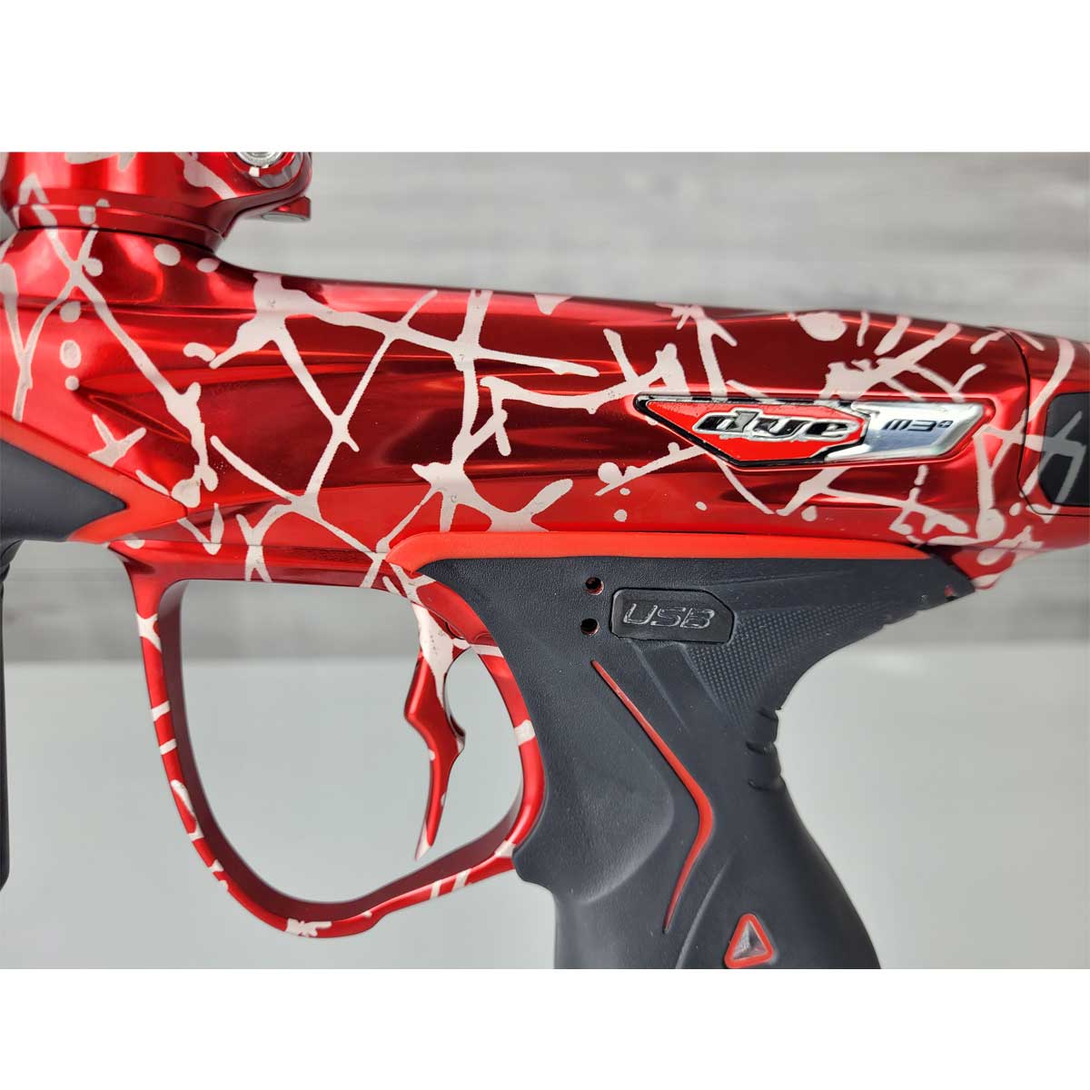 Close up of USB port cover USED Red and black with white splash pattern Dye M3+ Paintball Marker Red Splash on a woodgrain background.