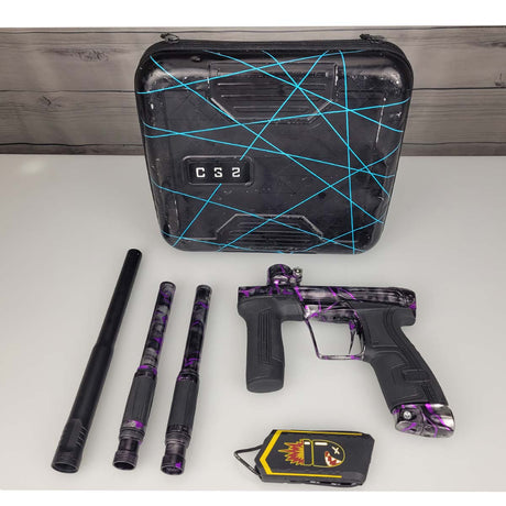 A USED Planet Eclipse CS2 Paintball Marker Black Purple Grey Splash on a woodgrain background laid out on a white surface, including a black and purple camouflage paintball marker, three paintball barrel parts, a black hard case with blue line graphics, and a yellow-and-black signal jammer. The marker has a black body with purple and gray camouflage patterns.