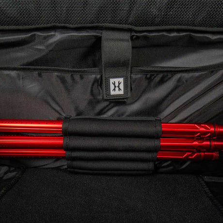 Close-up of a black soft-sided bag interior with a black strap and a small square logo patch, holding a bundle of bright red paintball barrels in black elastic loops.