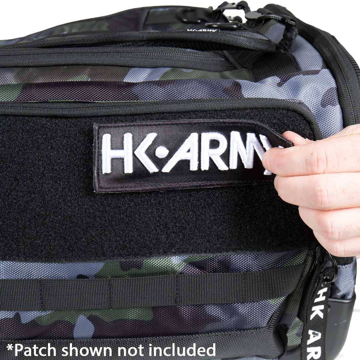 Close-up of a camouflage tactical bag with a black velcro patch holding a white HK-ARMY patch. A hand is adjusting the patch, and a text overlay at the bottom reads '*Patch shown not included'.