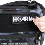 Close-up of a camouflage tactical bag with a black velcro patch holding a white HK-ARMY patch. A hand is adjusting the patch, and a text overlay at the bottom reads '*Patch shown not included'.