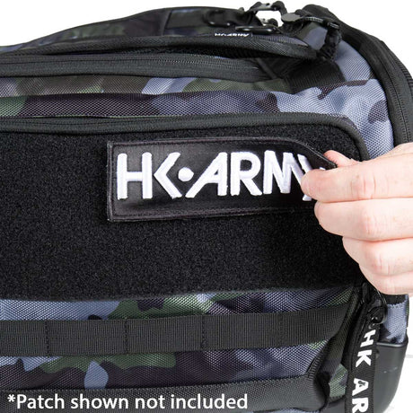 Close-up of a camouflage tactical bag with a black velcro patch holding a white HK-ARMY patch. A hand is adjusting the patch, and a text overlay at the bottom reads '*Patch shown not included'.