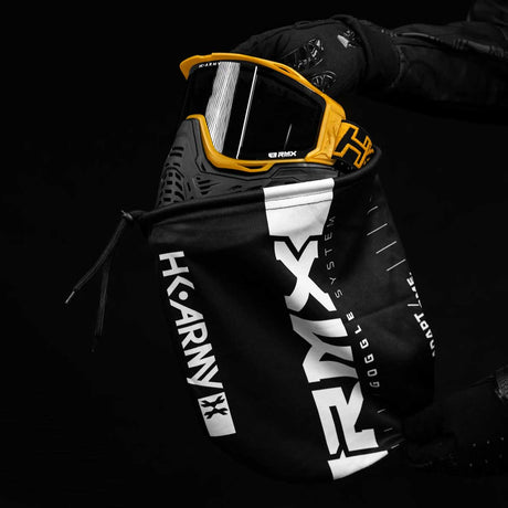 of person wearing HK Army RMX Thermal Paintball Goggles Black yellow Gold with smoke lens being placed inside a fabric goggle bag on a black background